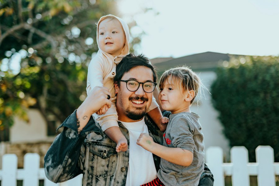 Smiling man in glasses carrying two young children outside a house